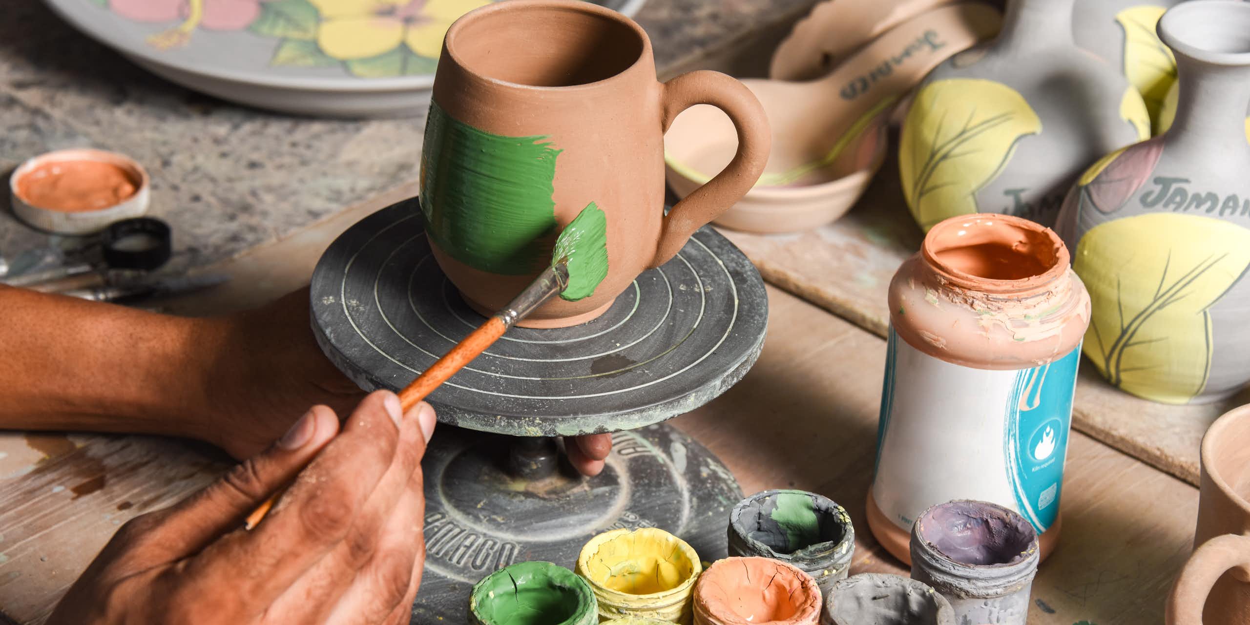 Potter's hands shown painting a clay mug, with other pots and painting accessories around it.