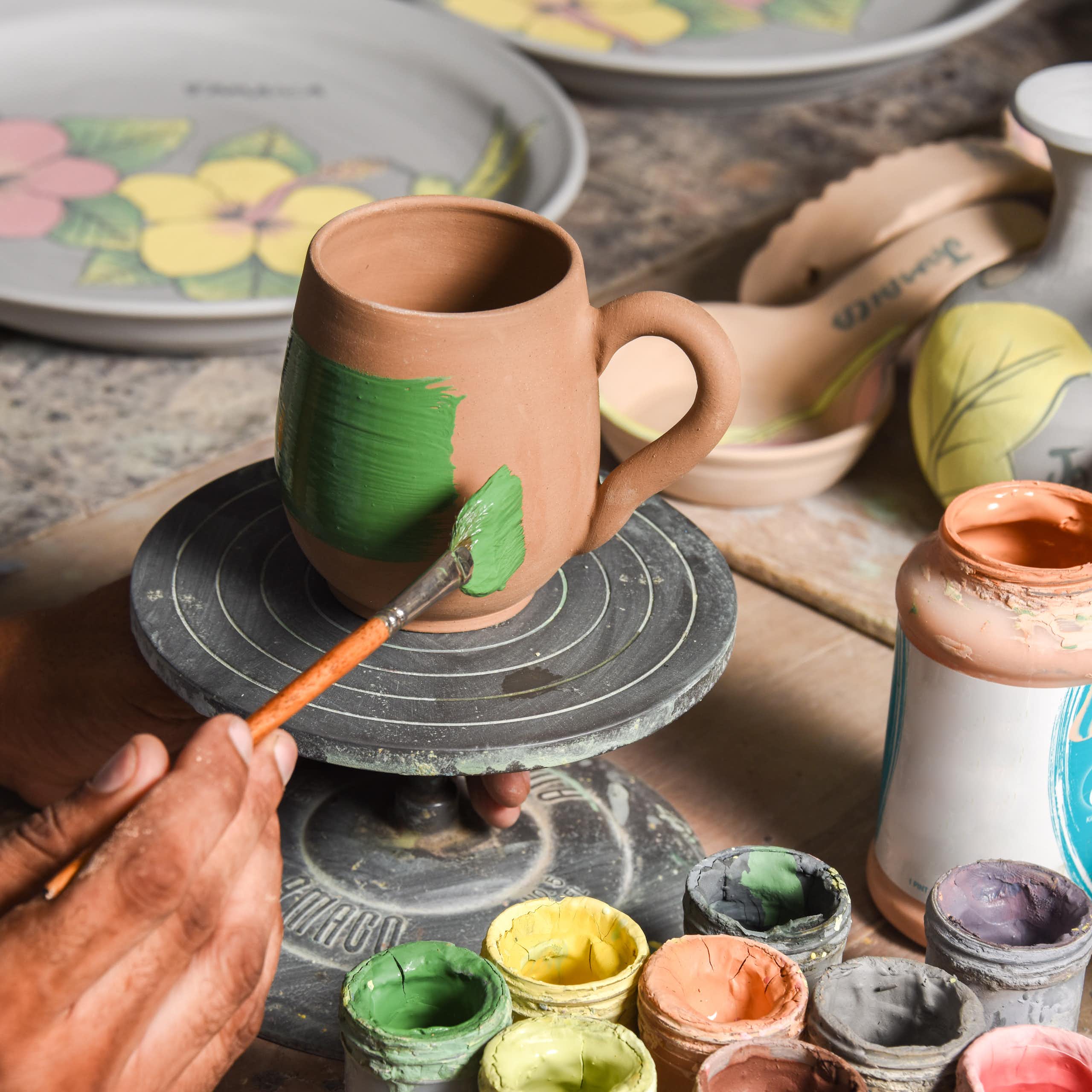Potter's hands shown painting a clay mug, with other pots and painting accessories around it.