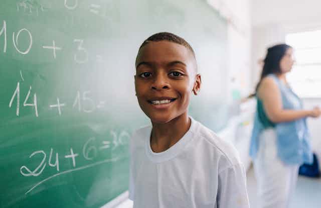 An African American boy smiles while at a chalkboard filled with math problems.