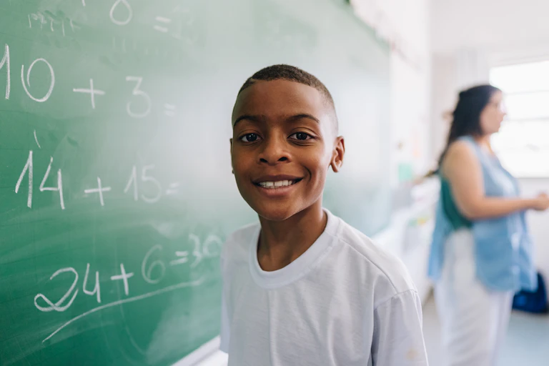 An African American boy smiles while at a chalkboard filled with math problems.