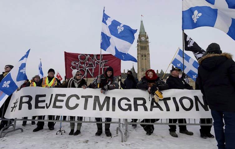 People carry a banner that reads Reject Migration with the Peace Tower behind them.