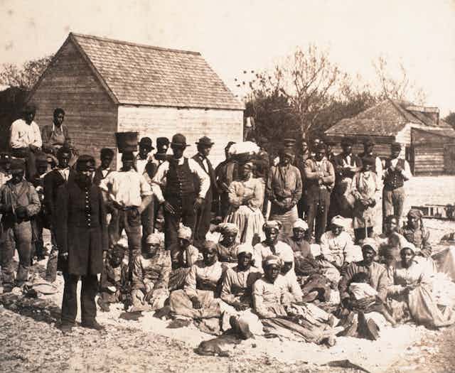 A group of Black people stand and sit in front of several small buildings. 