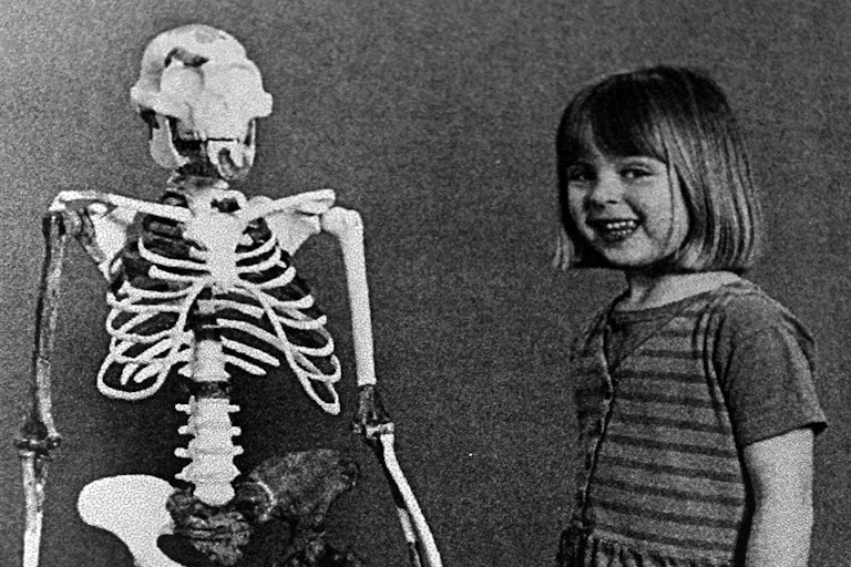 A smiling child, hands in her dress pockets, stands next to the reconstructed skeleton of an early humanoid. They are roughly the same height and have comparable head sizes.