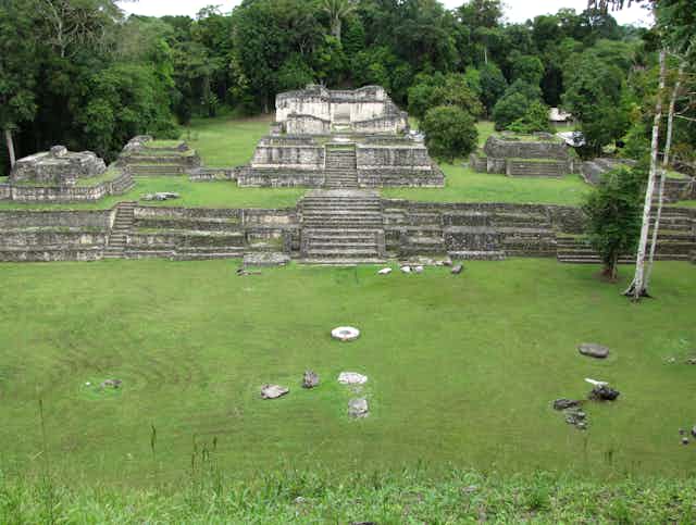 A cleared, grassy area with stone steps and ruins of monuments sits amid a forest.