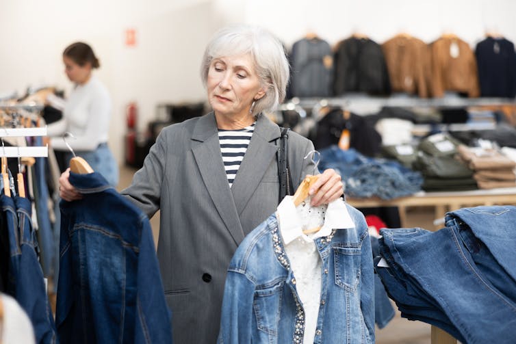 An older woman holds two hangers with denim jackets on them, one in each hand, while standing before a rack of clothes in a store