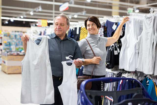 A middle-aged woman looks on as an elderly man holds up a hanger with a tank top on it