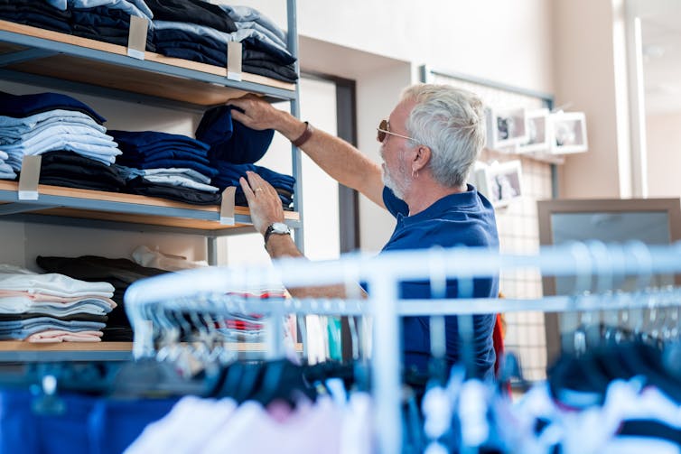A grey-haired man wearing sunglasses browses a stack of clothing on a shelf in a store