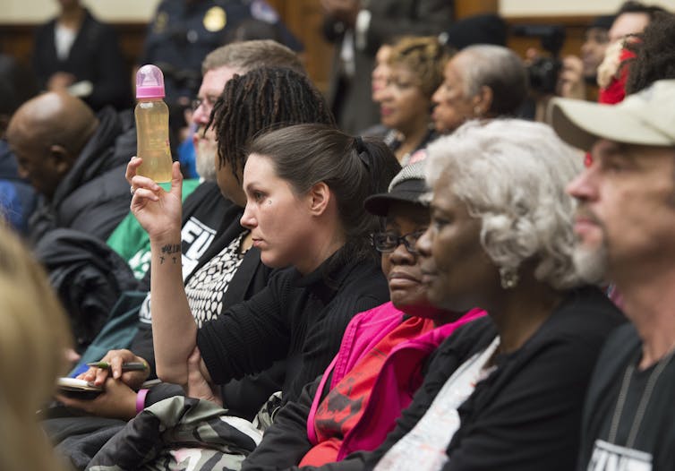 A woman holds a baby bottle with rust-colored water inside. The hearing room is packed with people around her.