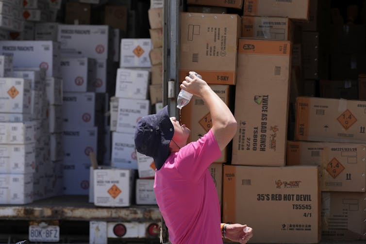 A man in a floppy hat and T-shirt pours water from a bottle into his mouth in front of a truck.
