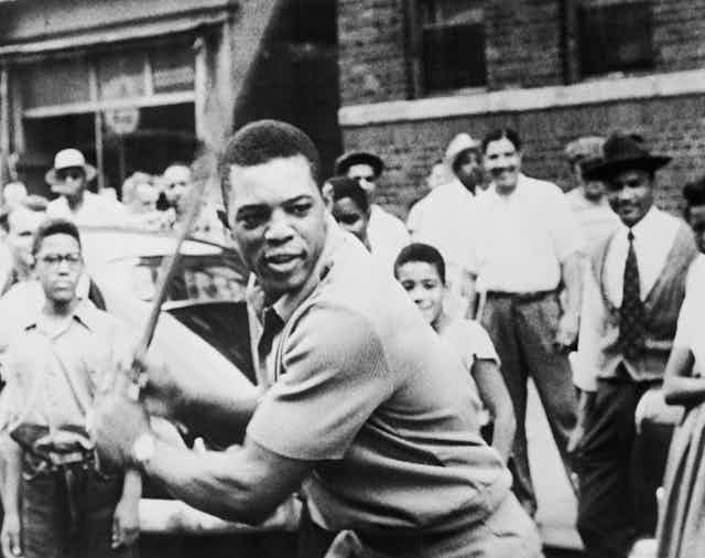 A Black man stands in front of a crowd with a baseball bat waiting for a pitch. 