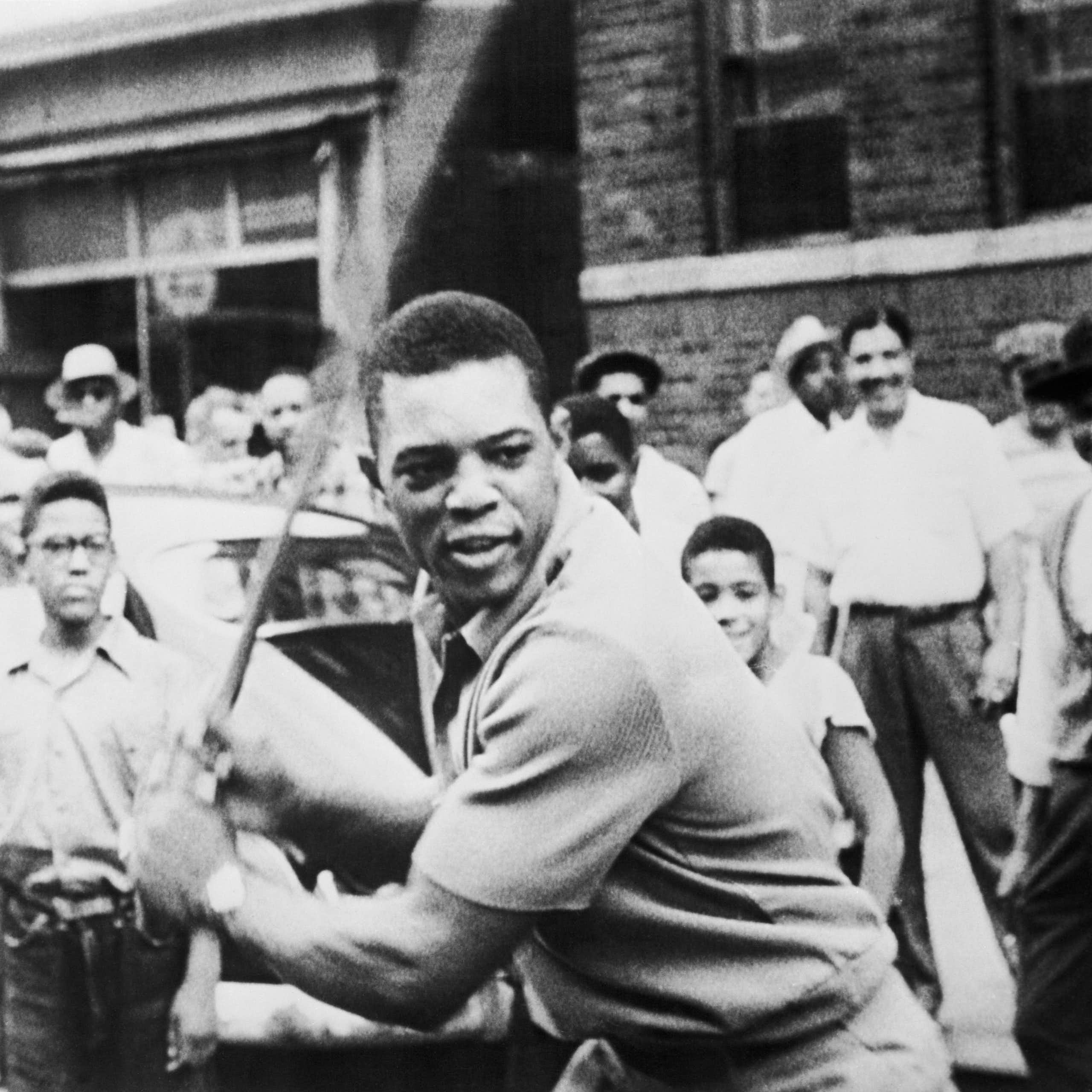 A Black man stands in front of a crowd with a baseball bat waiting for a pitch. 