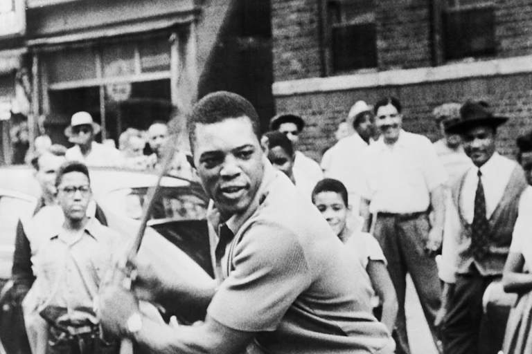 A Black man stands in front of a crowd with a baseball bat waiting for a pitch.