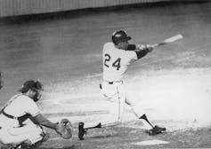 A Black man wearing baseball uniform stands at home plate and hits a homerun.