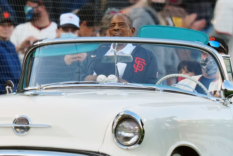 An elderly man in a vintage convertible looks out over a crowd.