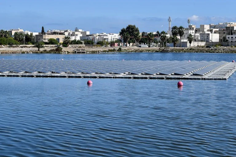 Des dizaines de lignes de panneaux solaires flottent sur un lac devant une scène tunisienne de bâtiments traditionnels blanchis à la chaux.