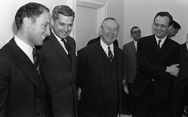 A black-and-white photo shows an older man standing with three younger men. All wear dark suits and ties.
