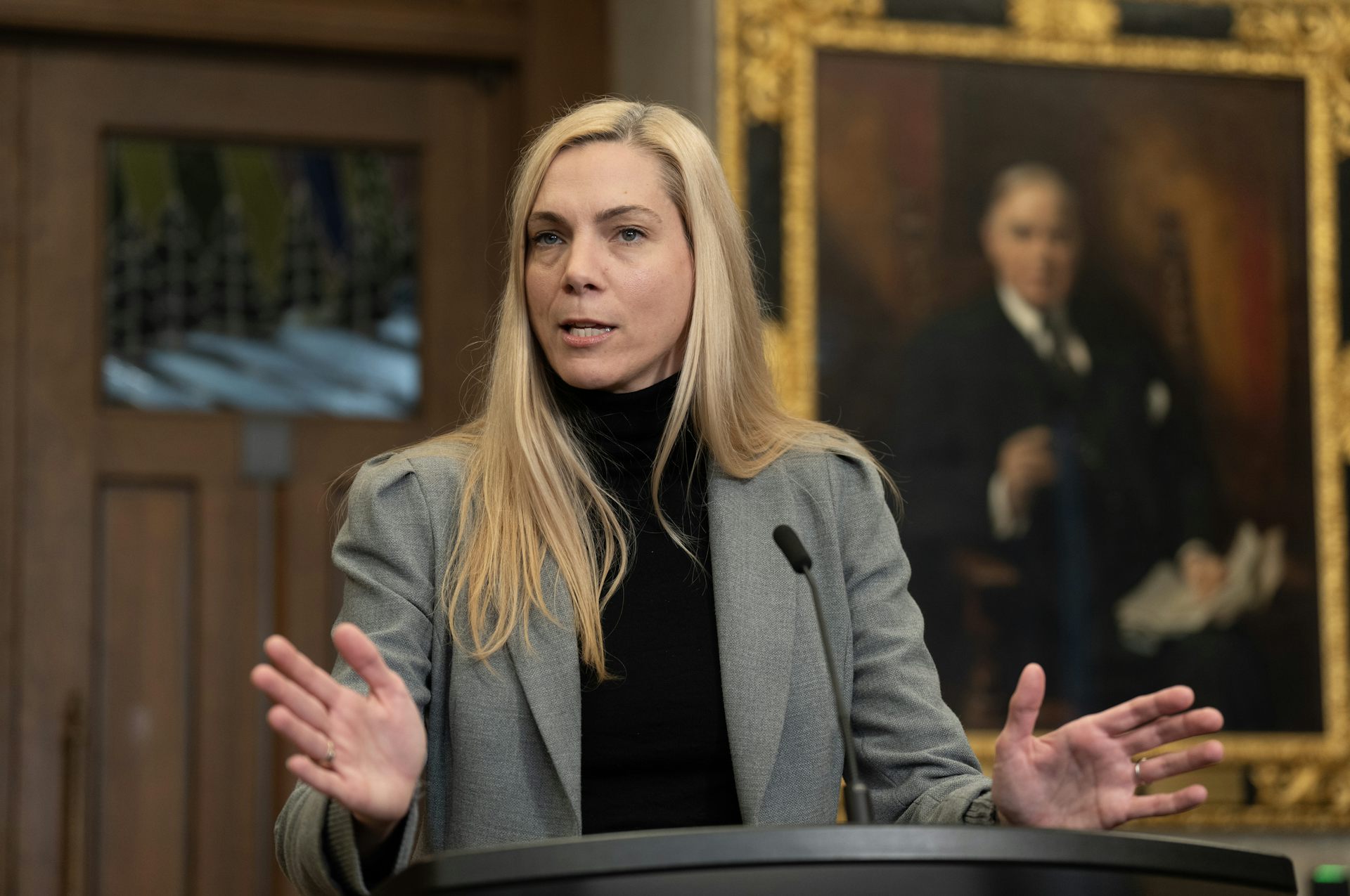 A middle-aged white woman with long blonde hair wearing a grey blazer speaks to people off-camera