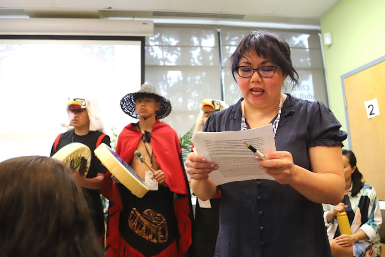 A woman in glasses speaks at an event with men in Indigenous garb standing behind her.