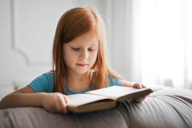 A young girl lies on a bed, reading a book.