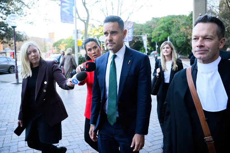A man in a suit and green tie walking outside, surrounded by his lawyers.