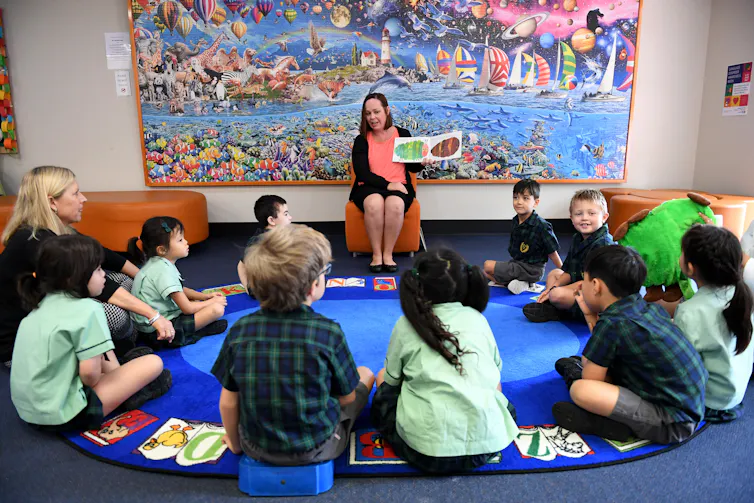A teacher sits on a chair in front of young students, who sit in a circle. The teacher holds an open book.