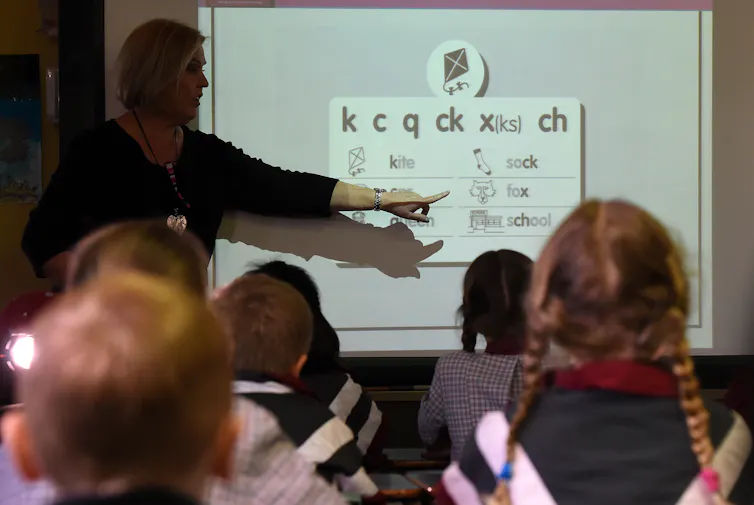 A teacher stands at a white board, pointing out sounds for 'k,' 'c' and 'q' in words. Students sit at desks watching.