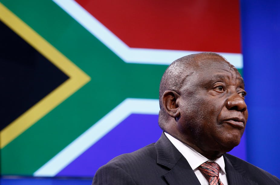Head and shoulders of a man in suit and tie, in front of the South African flag.