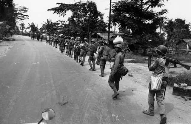 A black and white image of a long row of soldiers walking single file down a road, one looking back at the camera.