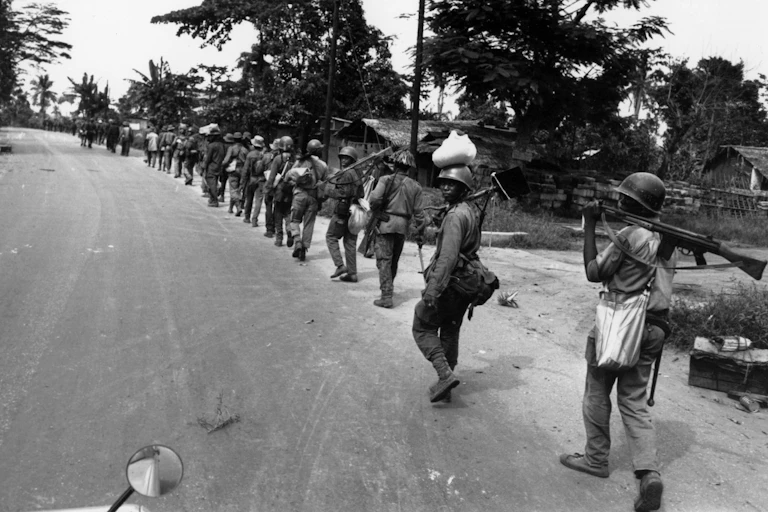 A black and white image of a long row of soldiers walking single file down a road, one looking back at the camera.