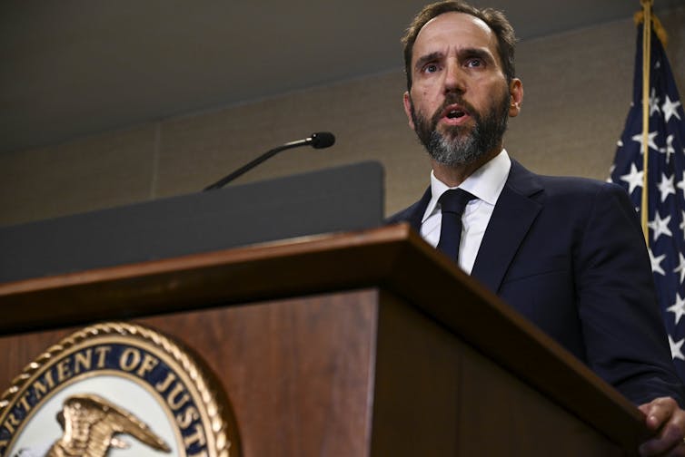 Un hombre de cabello oscuro, con barba y vestido con traje, habla en un atril que lleva el sello del Departamento de Justicia de Estados Unidos.