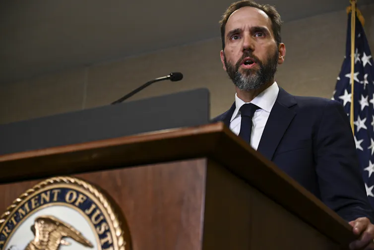 Um homem de cabelos escuros, com barba e vestindo um terno fala em um púlpito com o selo do Departamento de Justiça dos EUA.