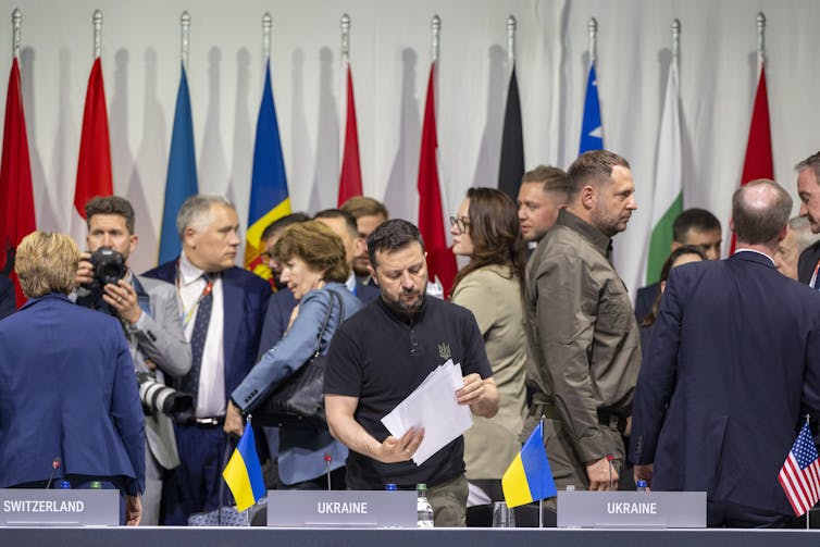 Ukrainian president Volodymyr Zelensky stands holding a sheaf of papers as delegates to the Ukraine peace conference mill around in different directions.