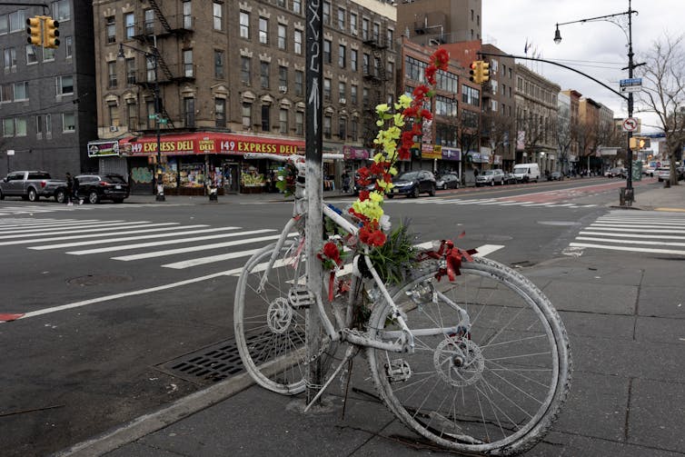 A bicycle, painted white and decorated with flowers, attached to a street pole at an urban intersection.