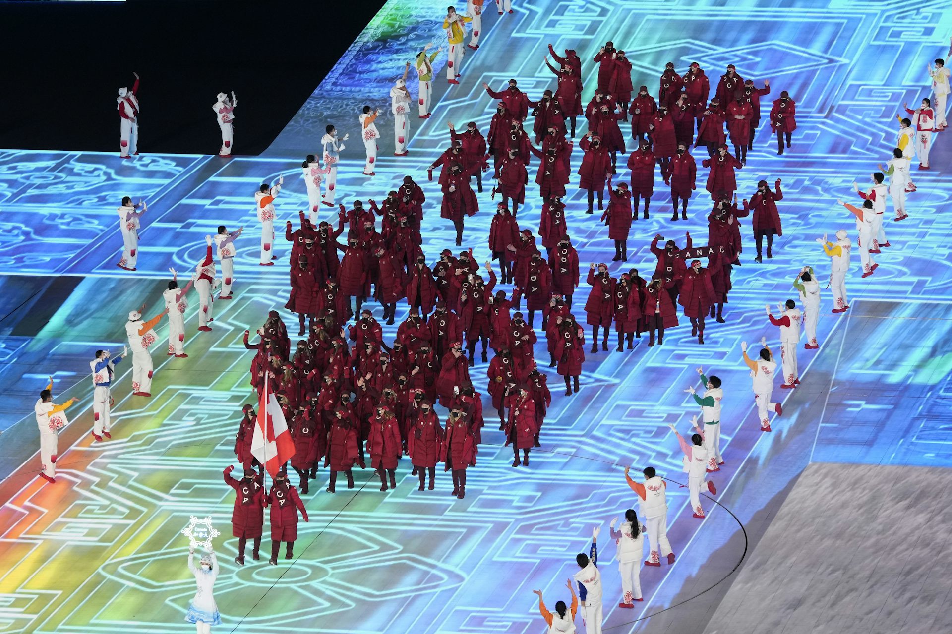 A crowd of people wearing red puffer coats follow two people holding a Canadian flag down a large, lit-up stage