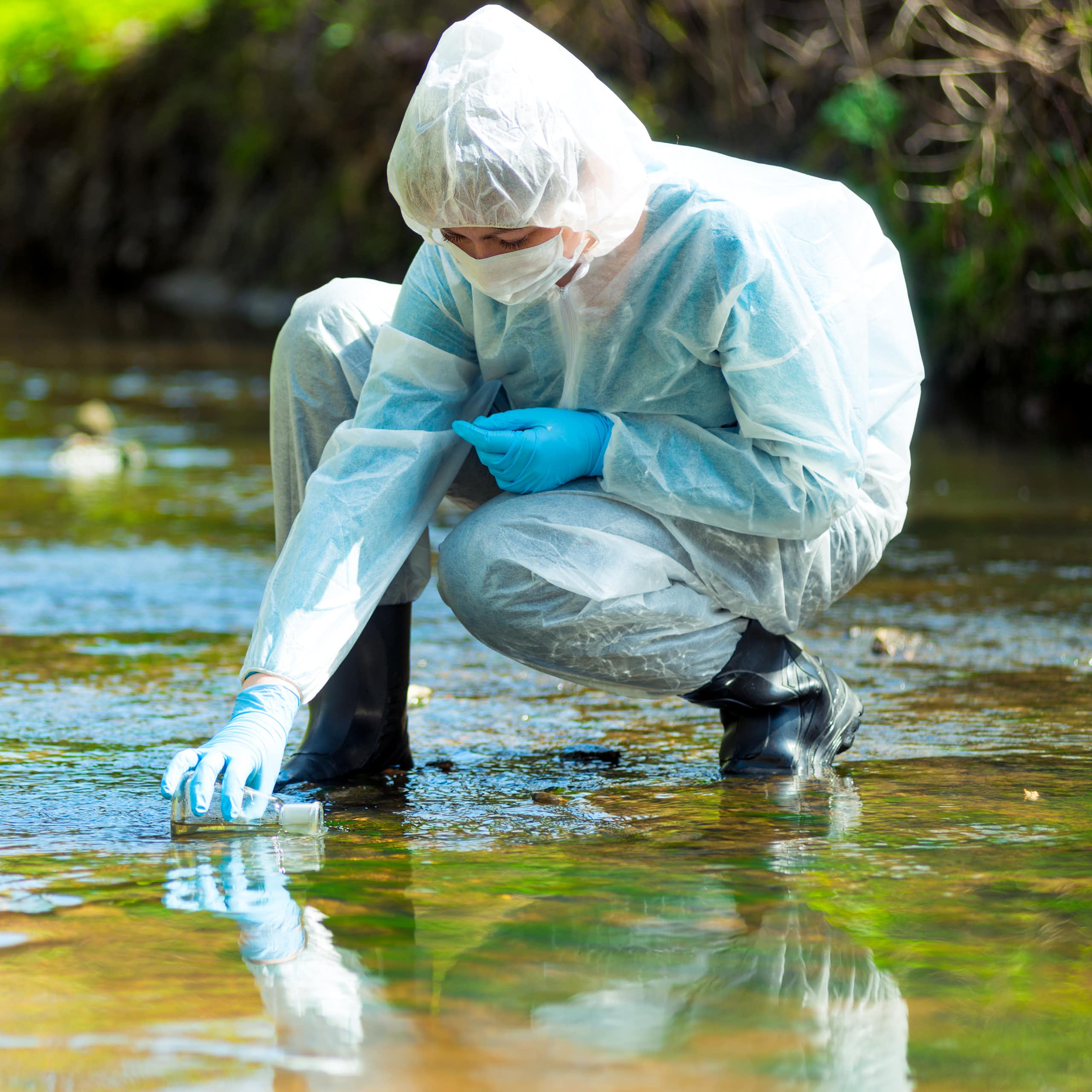 Scientist in PPE kneels down in river to collect water sample