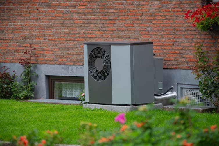 Grey air-source heat pump unit on old red brick house exterior, green grass and flowers in foregground