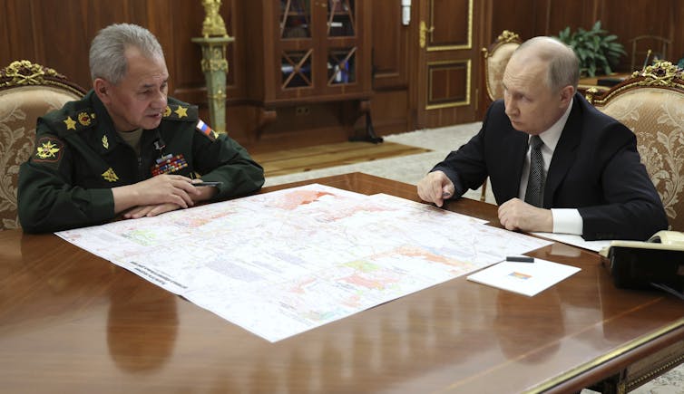 Two men sit at a wooden desk in an office talking, a large document on the desktop between them.