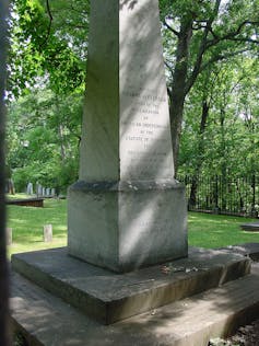 In a grassy area with trees there is an obelisk-shaped tomb.