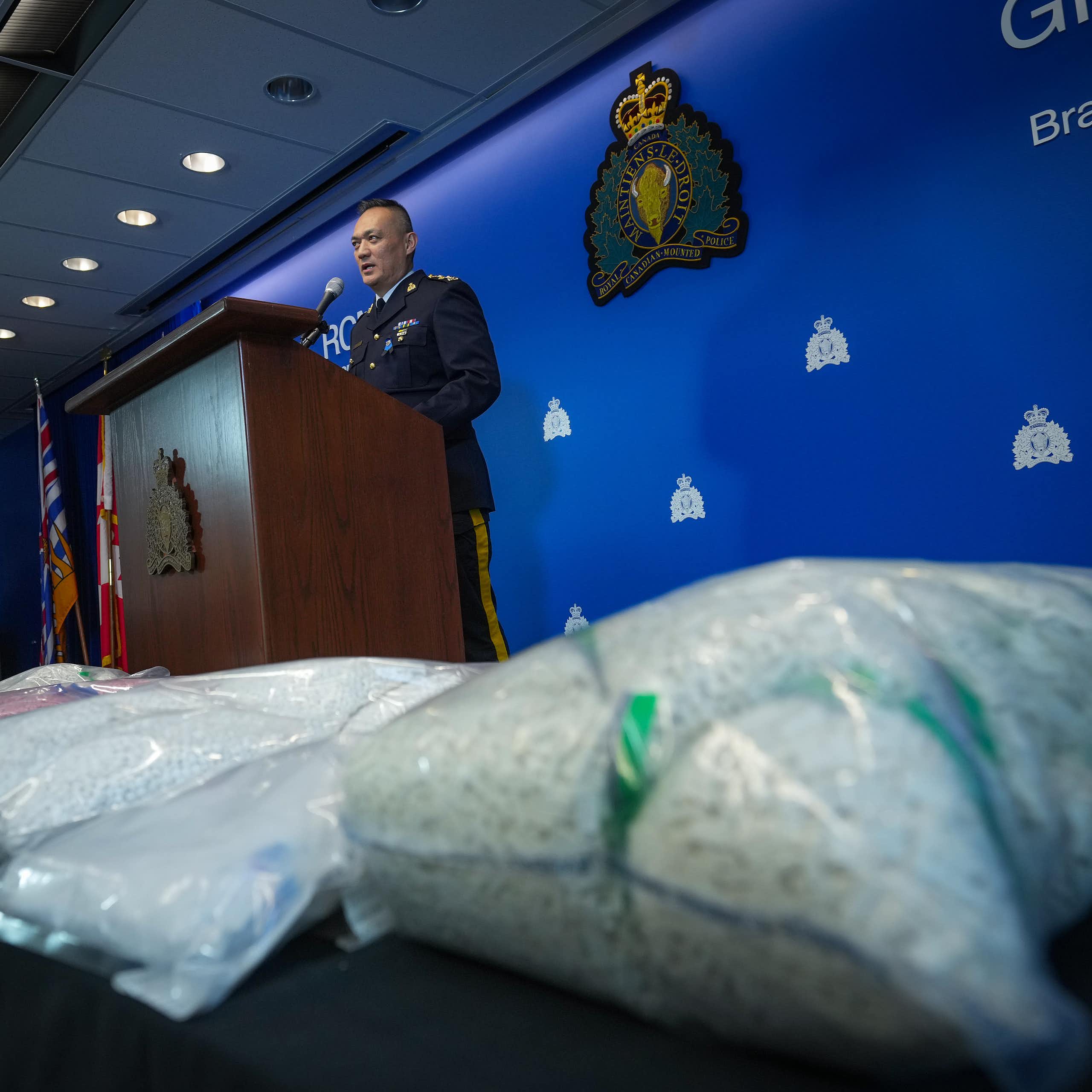 A police officer speaks into a microphone from behind a wooden lectern. Large packages of pills are in the foreground.