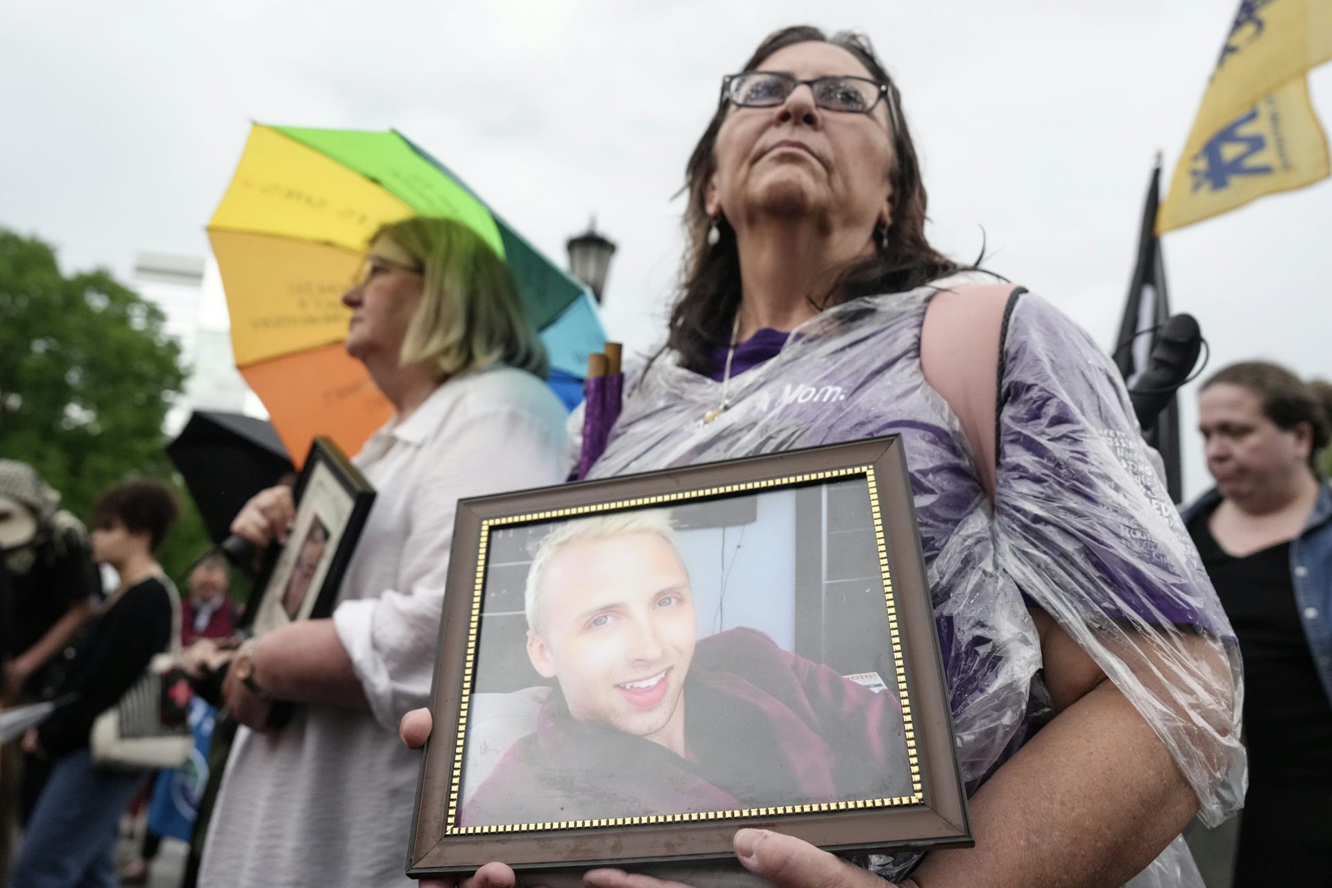 A woman stands in the rain holding a framed photo of a young blonde man.