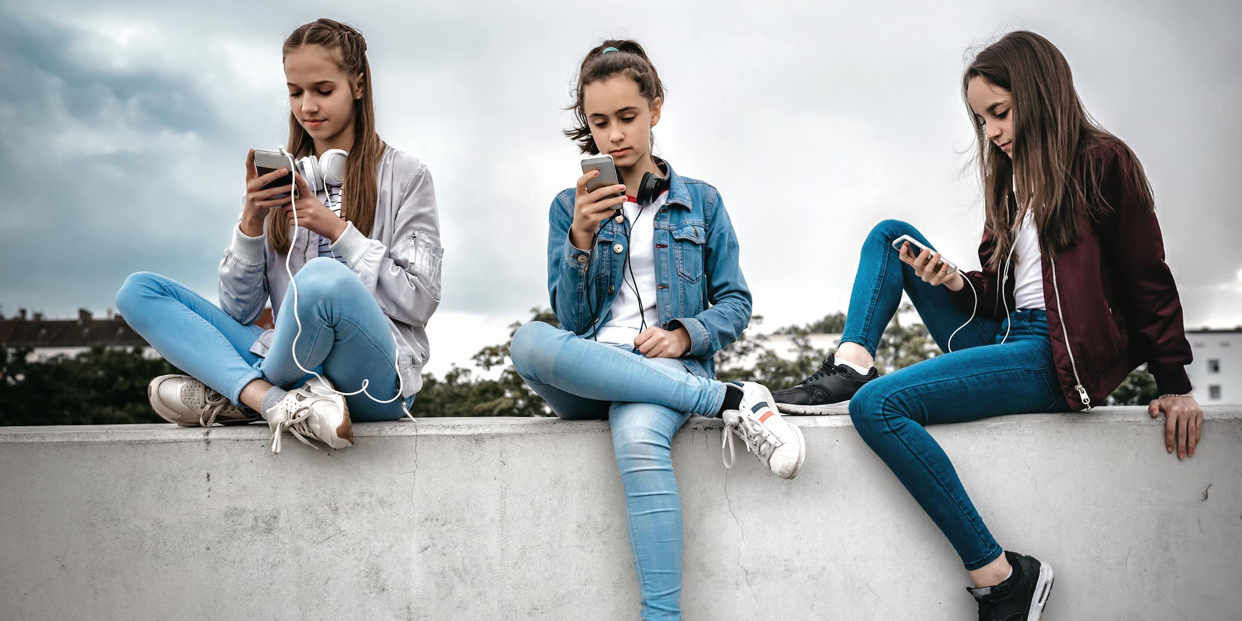 Three teenage girls sitting outdoors on concrete wall and looking at their smartphones.