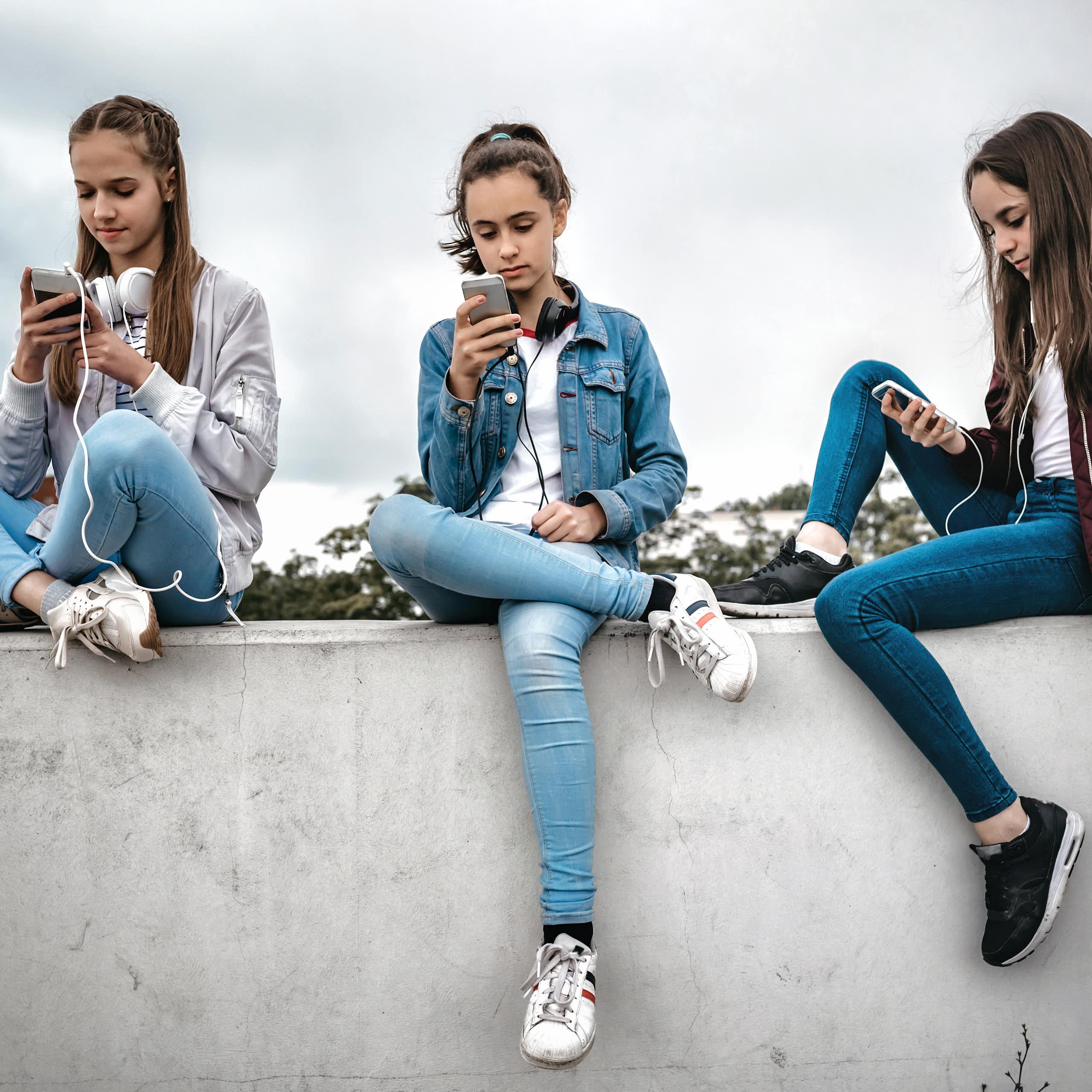 Three teenage girls sitting outdoors on concrete wall and looking at their smartphones.