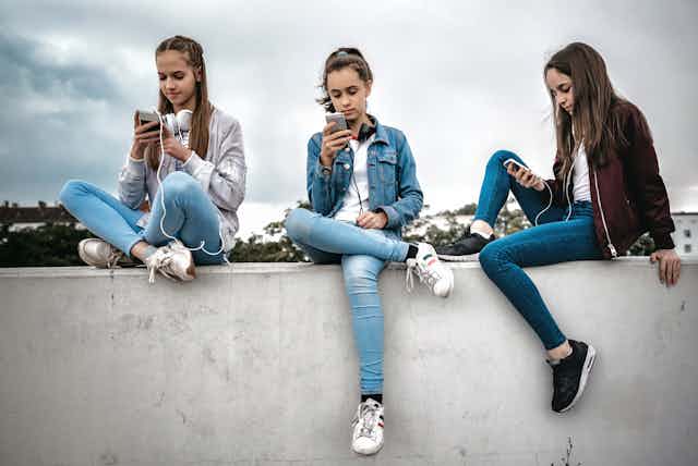 Three teenage girls sitting outdoors on concrete wall and looking at their smartphones.