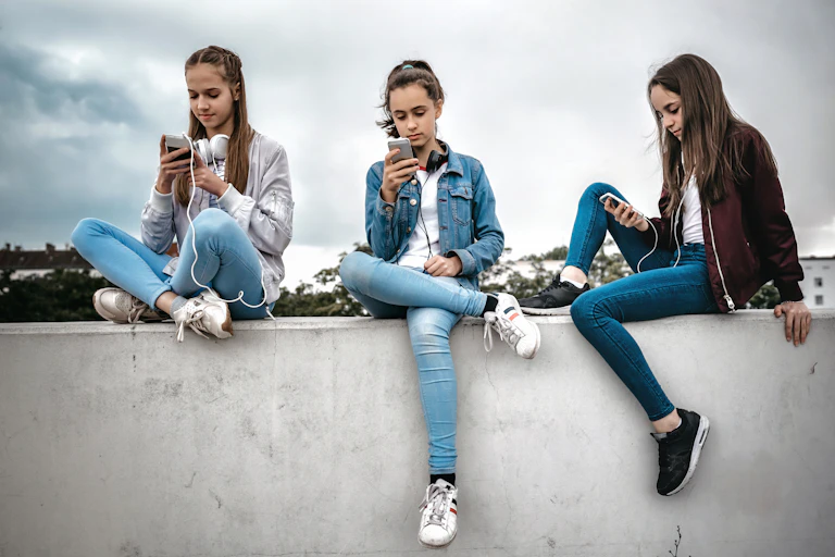 Three teenage girls sitting outdoors on concrete wall and looking at their smartphones.