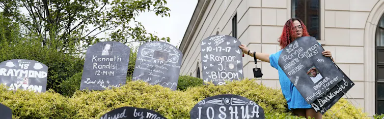 A woman stands among cardboard gravestones with the names of victims of opioid abuse.
