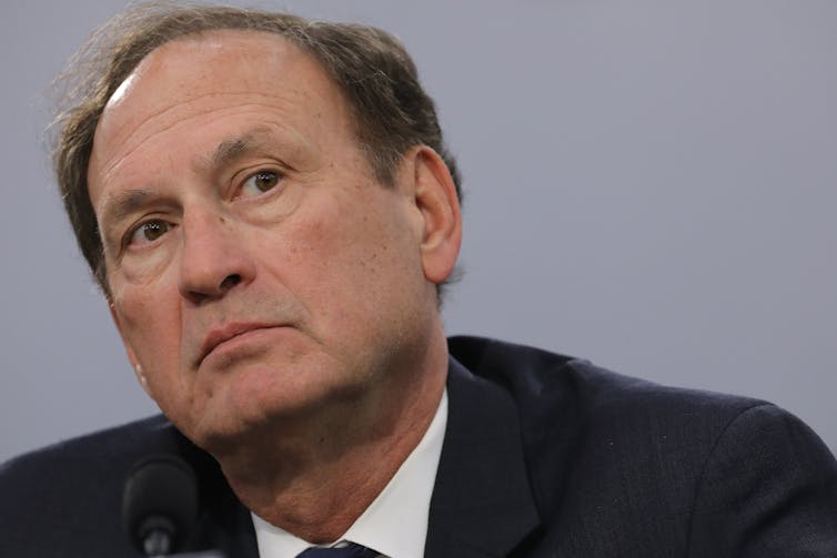 A dark-haired man in a suit cocks his head sideways.