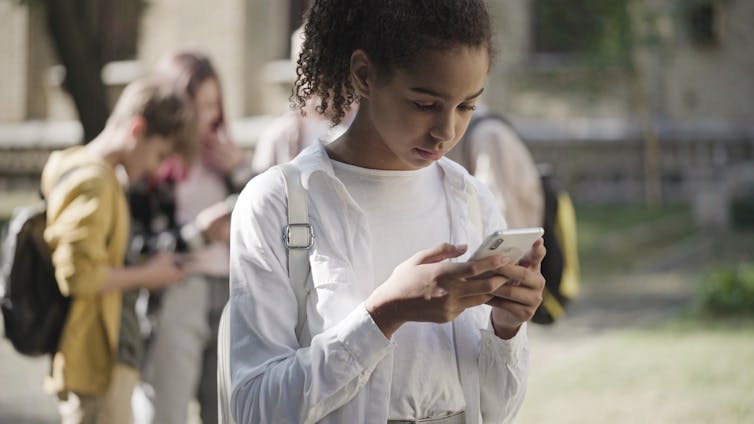 A girl looking at a smartphone with a cluster of other children with phones in the background