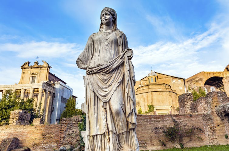 A statue of a vestal virgin is set against a blue sky and some ancient buildings.