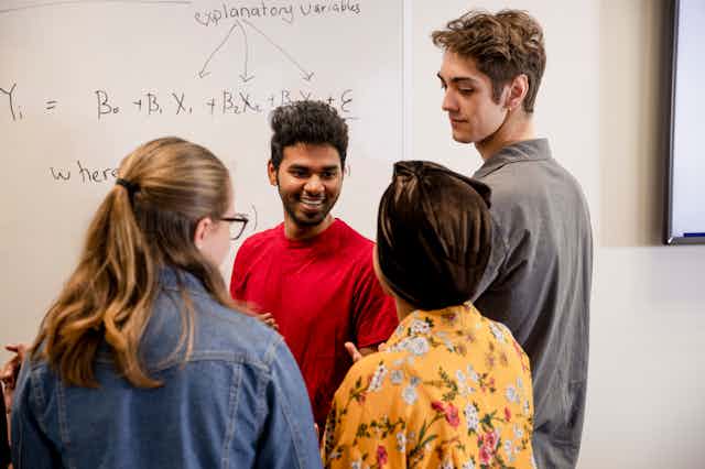 Four people stand in front on a white board, talking. An equation is written on the board. 