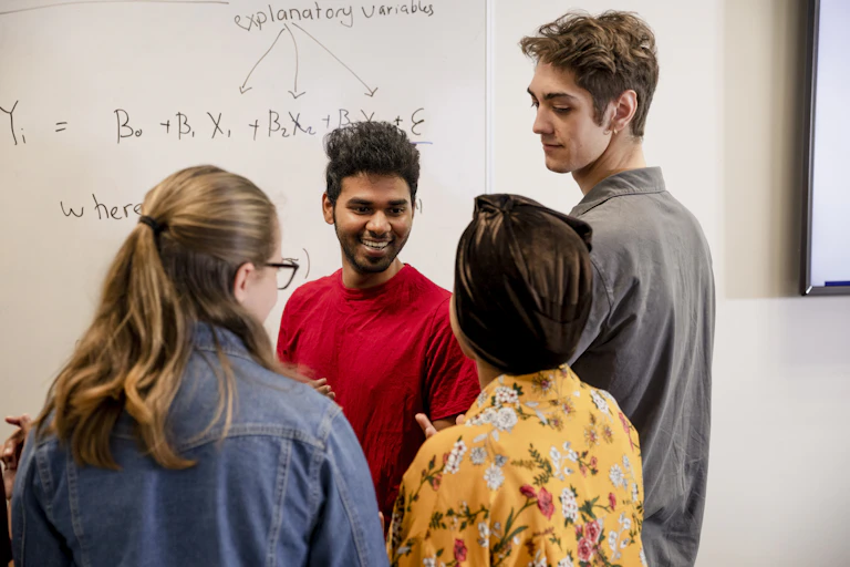Four people stand in front on a white board, talking. An equation is written on the board.